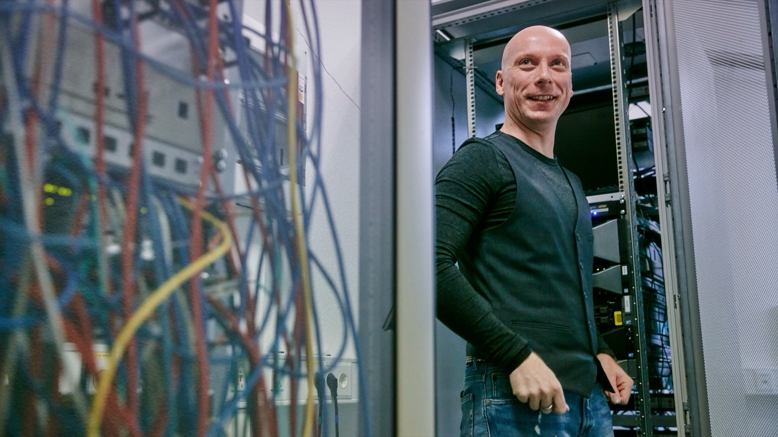 Philipp Brühl lächelt neben einem Serverschrank, links daneben hängt ein wirres Bündel bunter Netzwerkkabel.